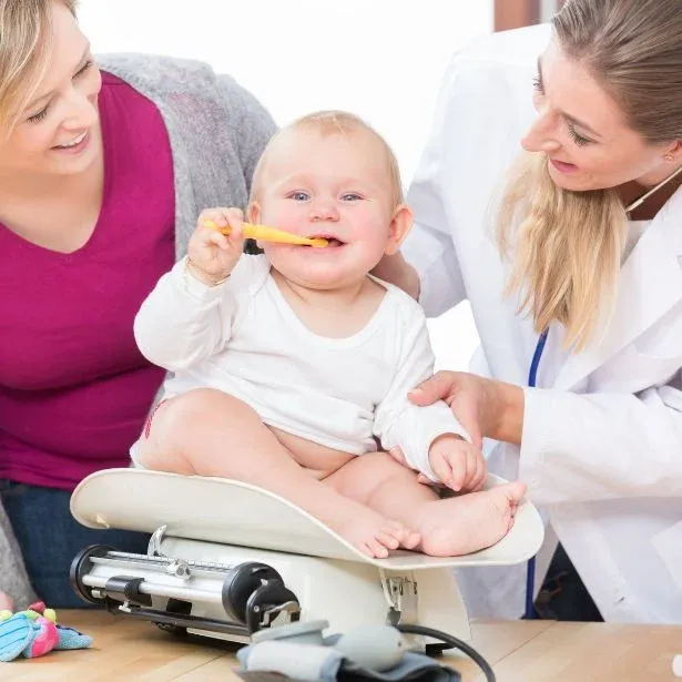Una donna ed una dentista che osservano un bimbo piccolo seduto su una bilancia